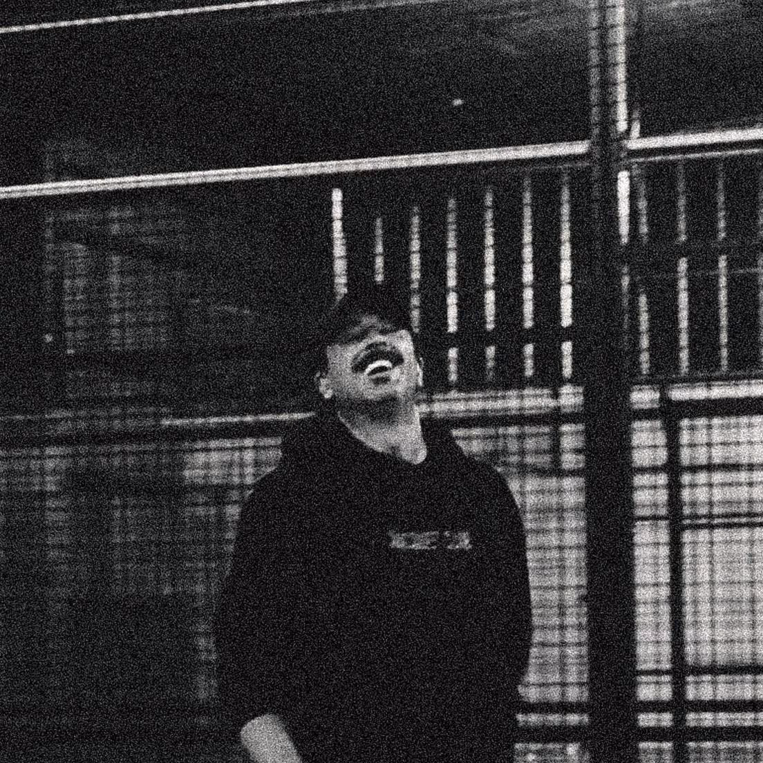Black-and-white grainy photo of a man wearing a cap and hoodie laughing with a fence in the background.