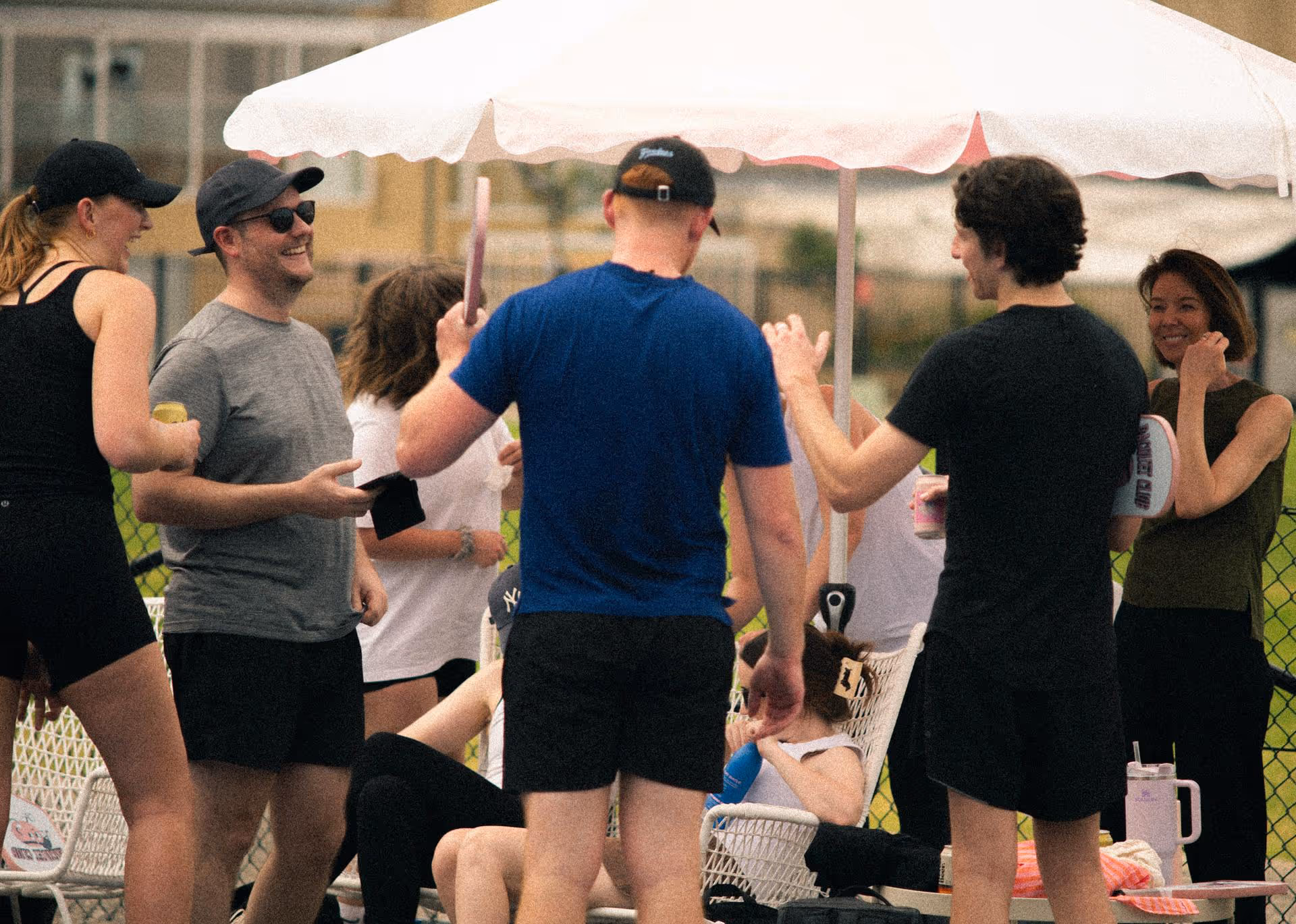 Group of people in casual sportswear socializing outdoors under a white umbrella, some holding pickleball paddles and drinks.