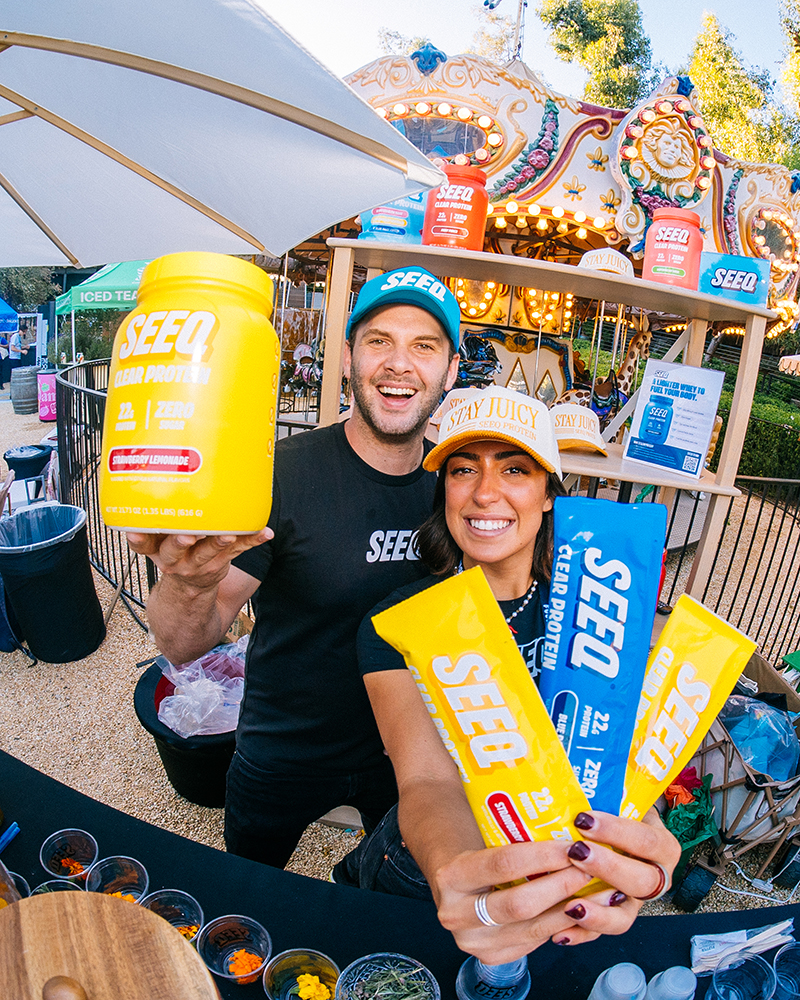Two smiling people wearing SEEQ hats and shirts holding SEEQ protein powder containers and bars at an outdoor event with a carousel in the background.