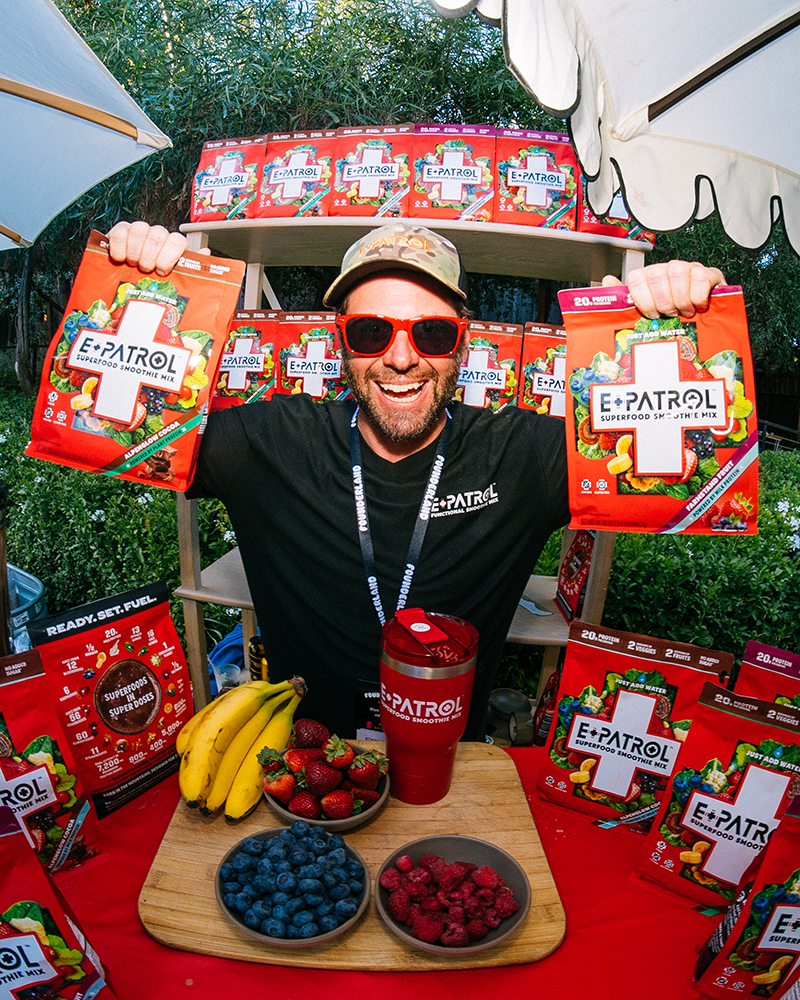 Man with red sunglasses and cap holding two bags of E-Patrol superfood smoothie mix at a booth with fresh fruit and product display.