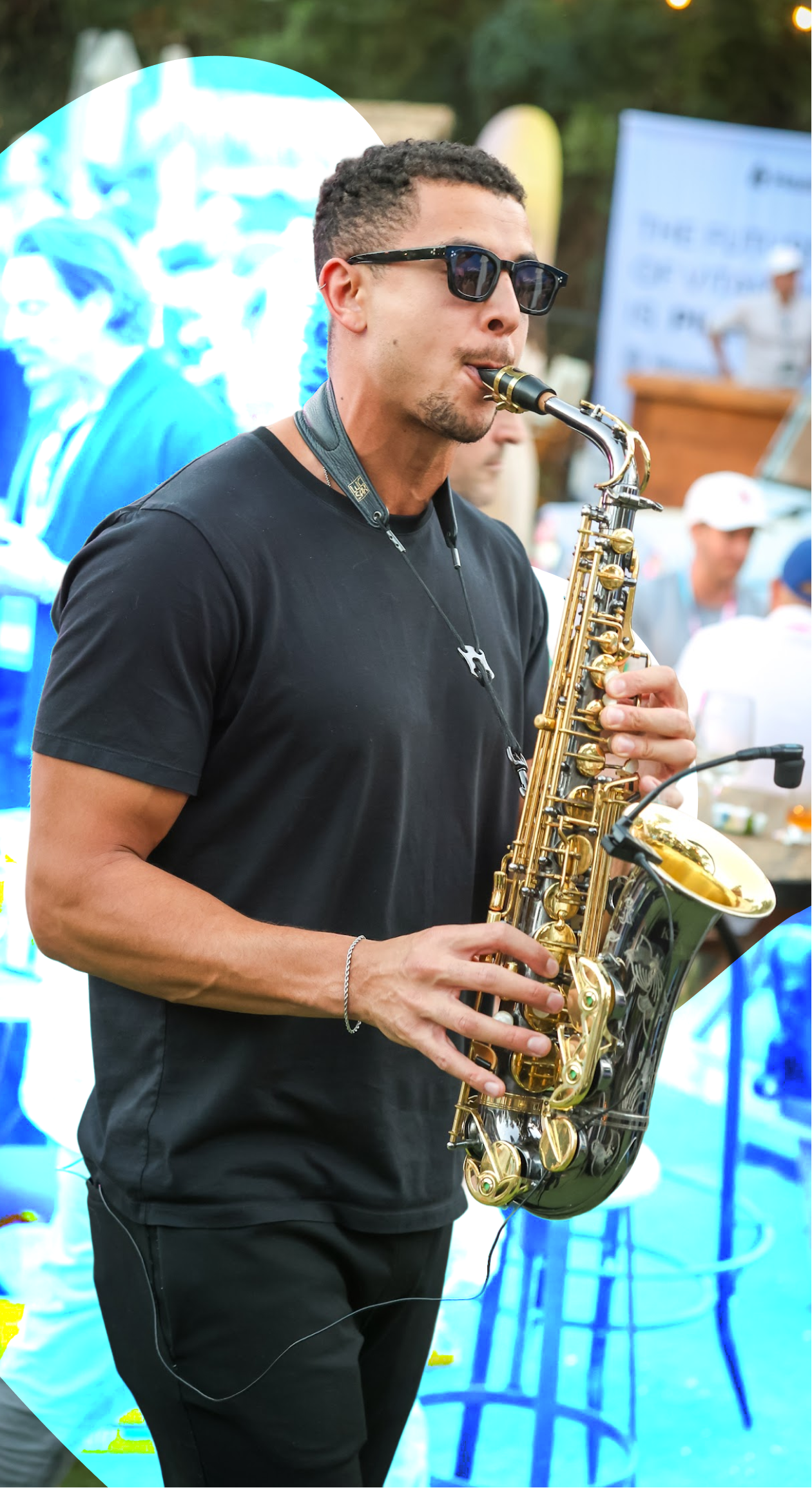 Man wearing sunglasses playing a gold and black saxophone at an outdoor event.