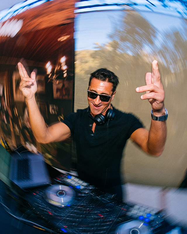 DJ wearing sunglasses and headphones, energetically mixing music at a DJ console with a laptop, with a blurred swirling motion effect in the background.