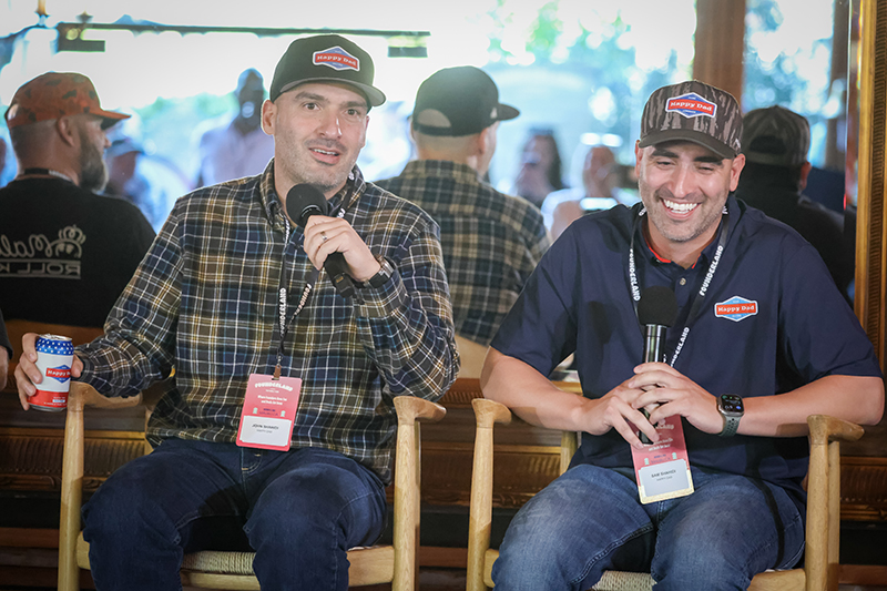 Two men wearing 'Happy Dad' hats and holding microphones, sitting on chairs and smiling during an event.