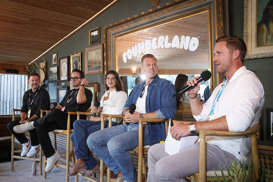Five people sitting on wooden chairs in a panel discussion setting with microphones, a large mirror behind them showing the word 'Founderland'.