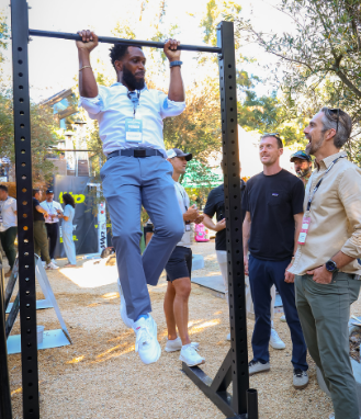 Man in business attire doing pull-ups on an outdoor exercise bar while three other men watch and smile.