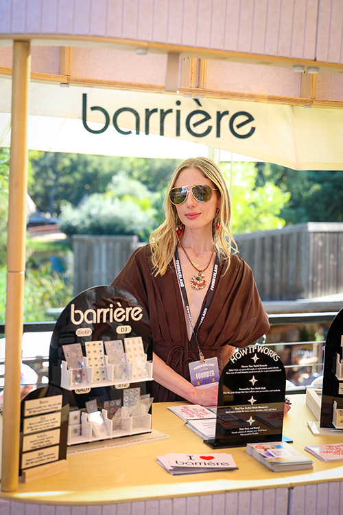 Woman wearing sunglasses and a brown dress standing behind a Barrière skincare booth displaying products and informational signs outdoors.