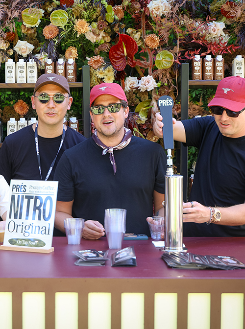 Three men wearing sunglasses and hats serving PRÉS Nitro Original protein coffee at an outdoor stall with floral decorations.