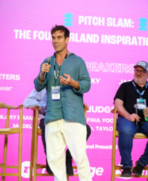 Man in a blue shirt and white pants speaking on stage at the Pitch Slam event with pink background screen and seated judges behind him.