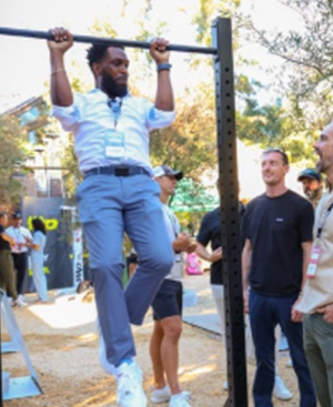 Man in business attire doing pull-ups on an outdoor exercise bar with others watching and smiling.