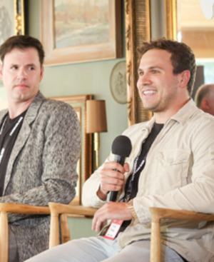 Two men seated indoors during a panel discussion, one holding a microphone and speaking, the other listening attentively.