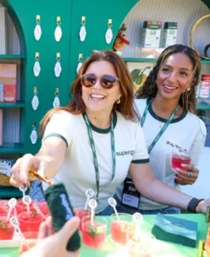 Two smiling women handing out small cups of red-colored drink at an event booth.