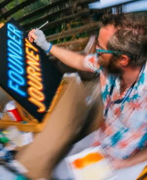 Man painting the words 'FOUNDER JOURNEY' in blue and orange on a black canvas outdoors.