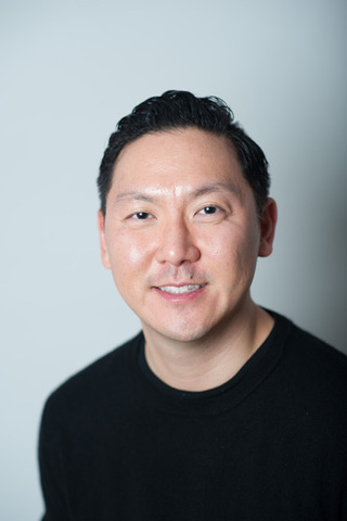Portrait of a smiling man with short black hair wearing a black sweater against a gray background.