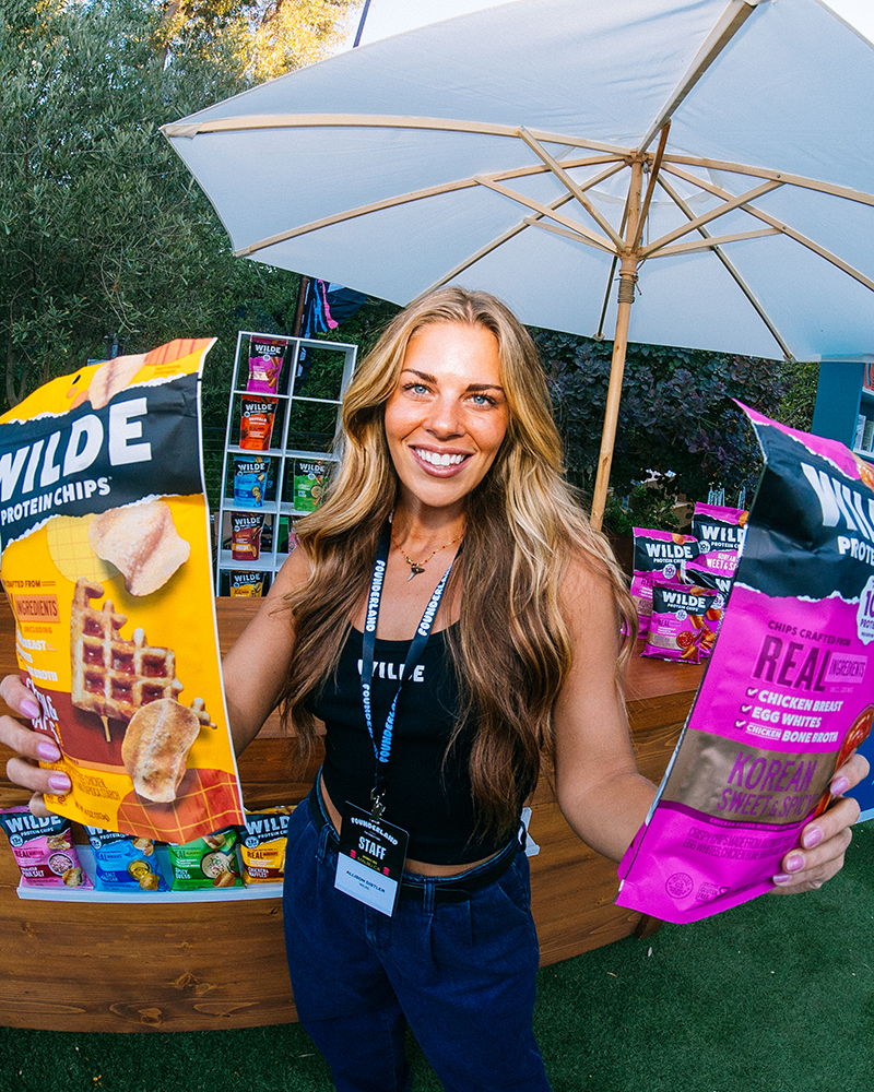 Smiling woman holding two large bags of Wilde protein chips, standing outdoors under a white umbrella at a display booth with more chip bags.