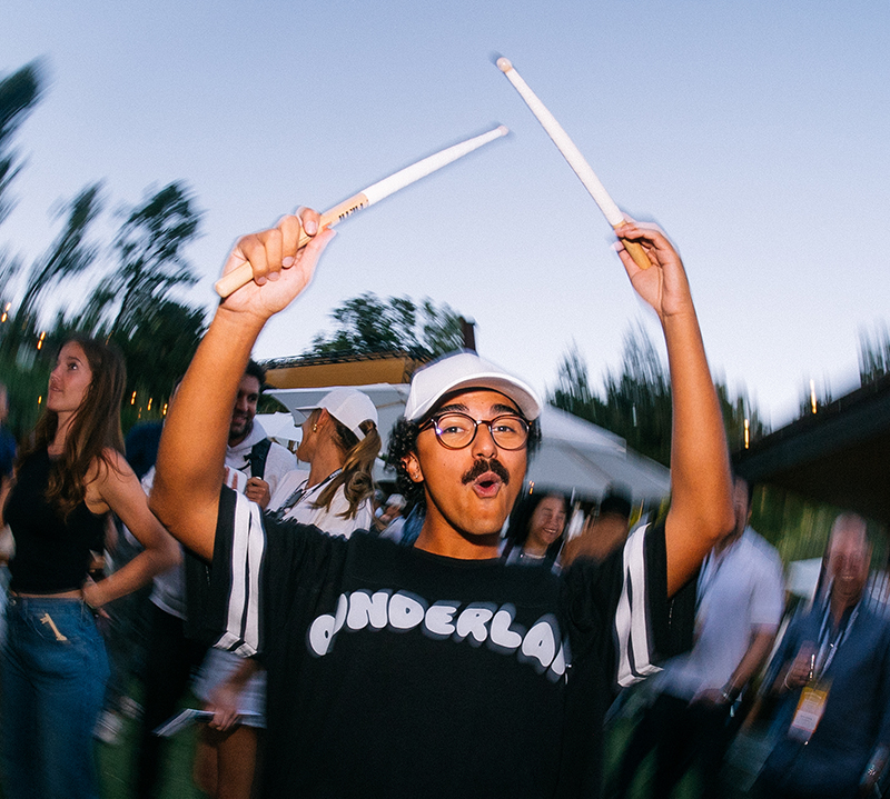 Young man wearing a white cap and glasses energetically holding drumsticks above his head at an outdoor event with people in the background.