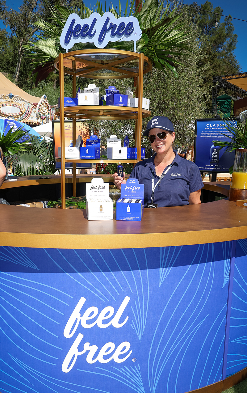 Smiling woman wearing sunglasses and a navy blue feel free cap and shirt, standing behind a blue and gold booth displaying feel free products outdoors.