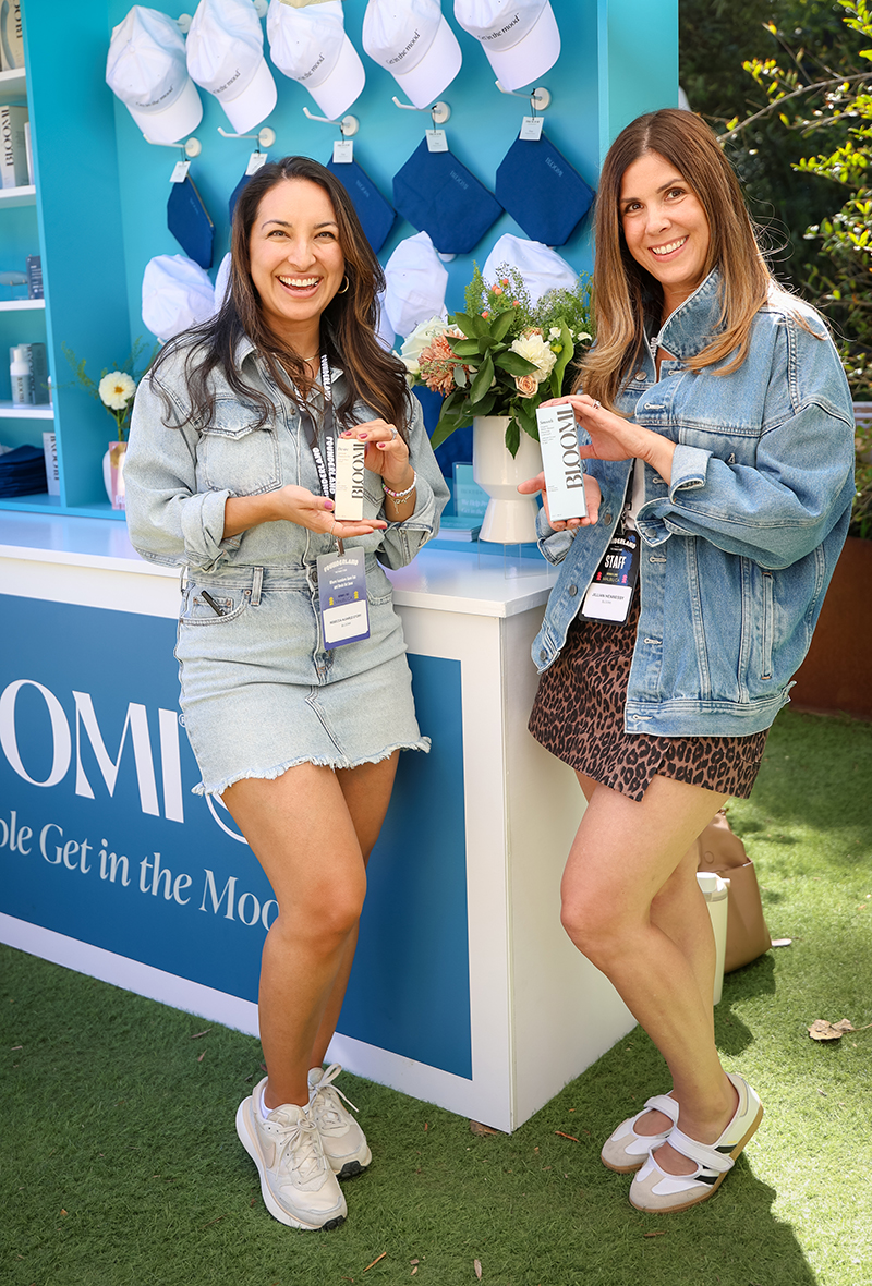 Two smiling women holding Bloomi skincare products, standing in front of a blue display with white caps and flowers.