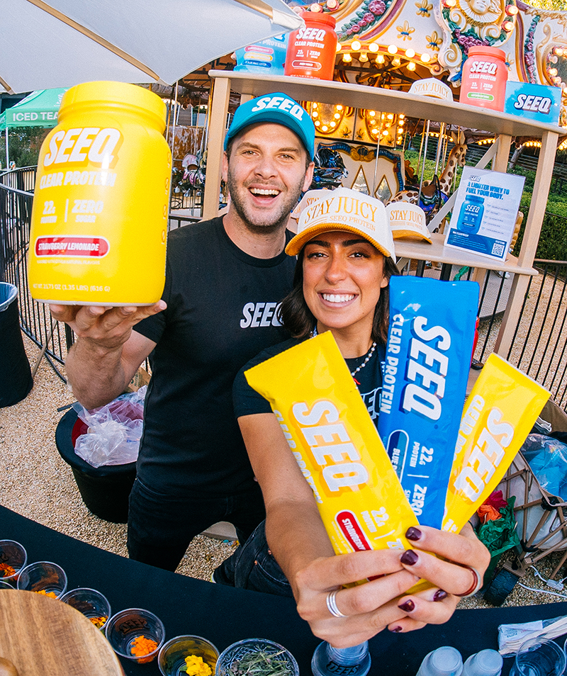 Two smiling people holding SEEQ Clear Protein products in front of a colorful outdoor booth.