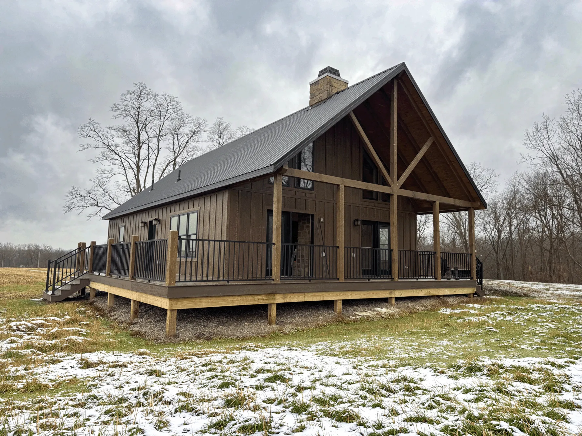 Rustic Amish-built modular log cabin home with a covered front porch, black metal roof, exposed timber framing, and stone chimney in a snowy Ohio countryside setting.