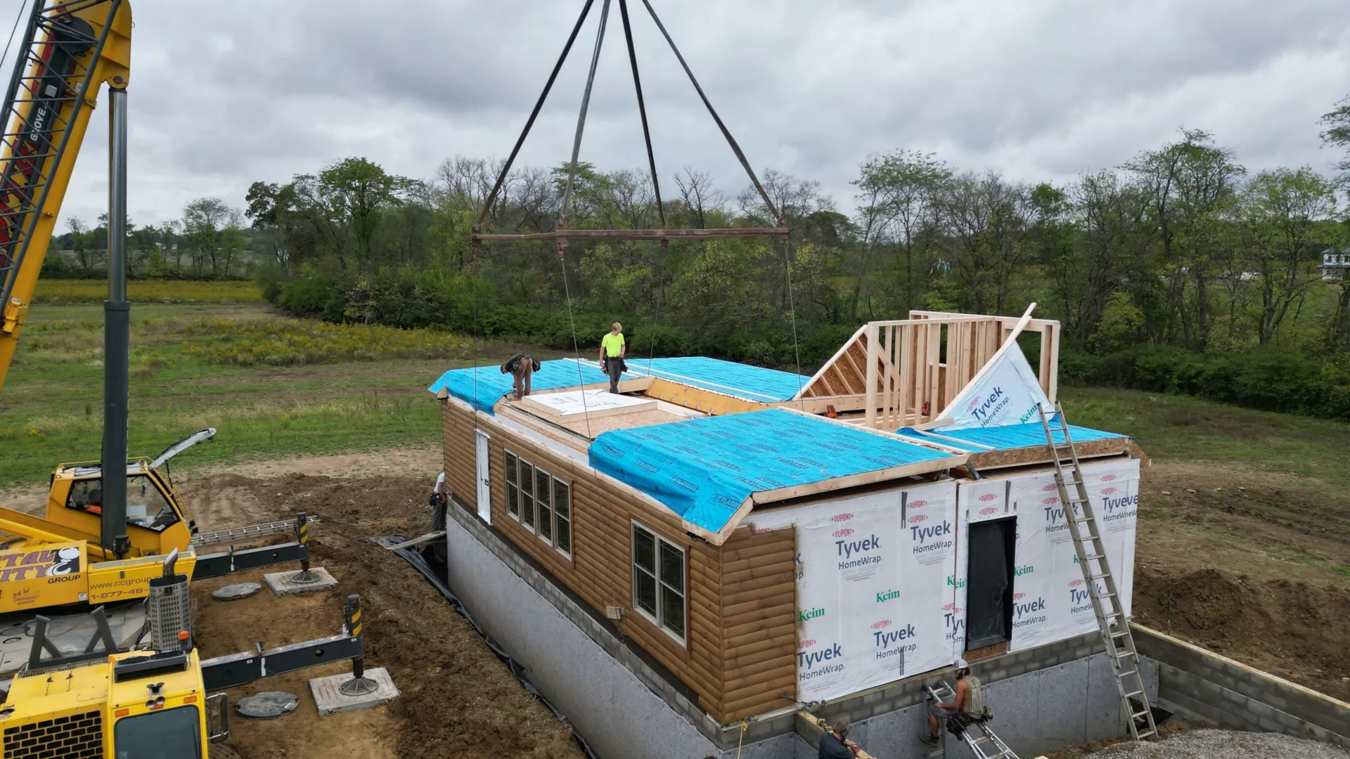 Modular log home being assembled on a foundation with a crane lifting a roof section into place during the modular home construction process.