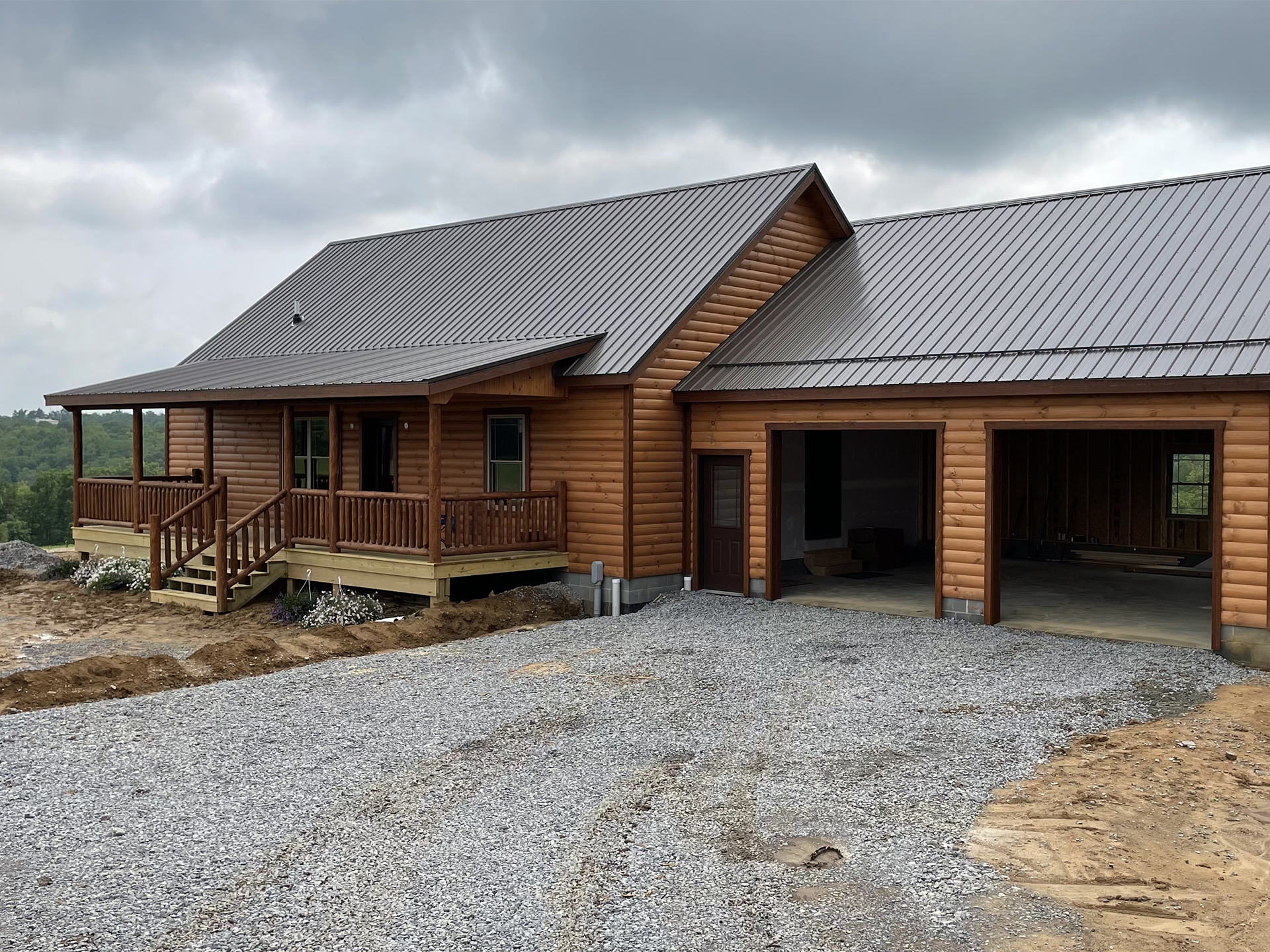 Modular log cabin home with log-style siding, a dark metal roof, a covered front porch with log railings, and an attached two-bay garage, set on a gravel drive overlooking wooded hills