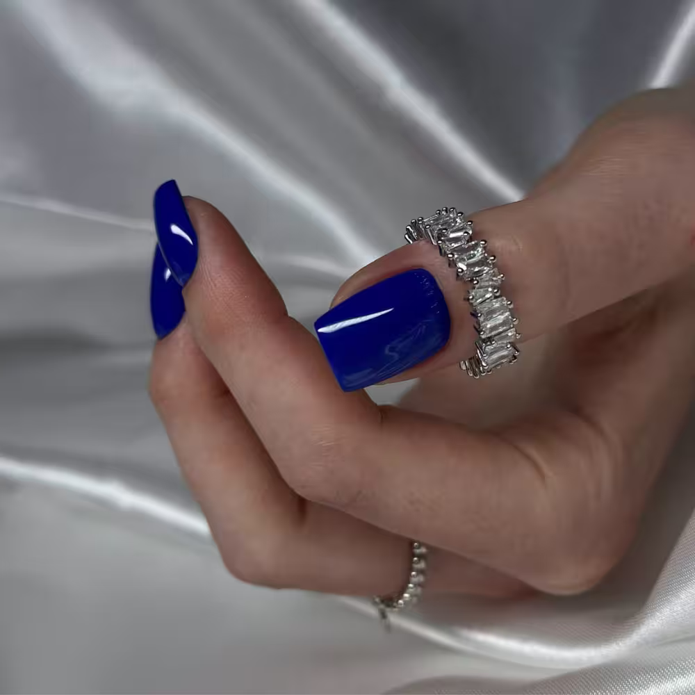 Hand with bright blue polished nails wearing a silver ring with clear rectangular stones, resting on a white silky fabric.