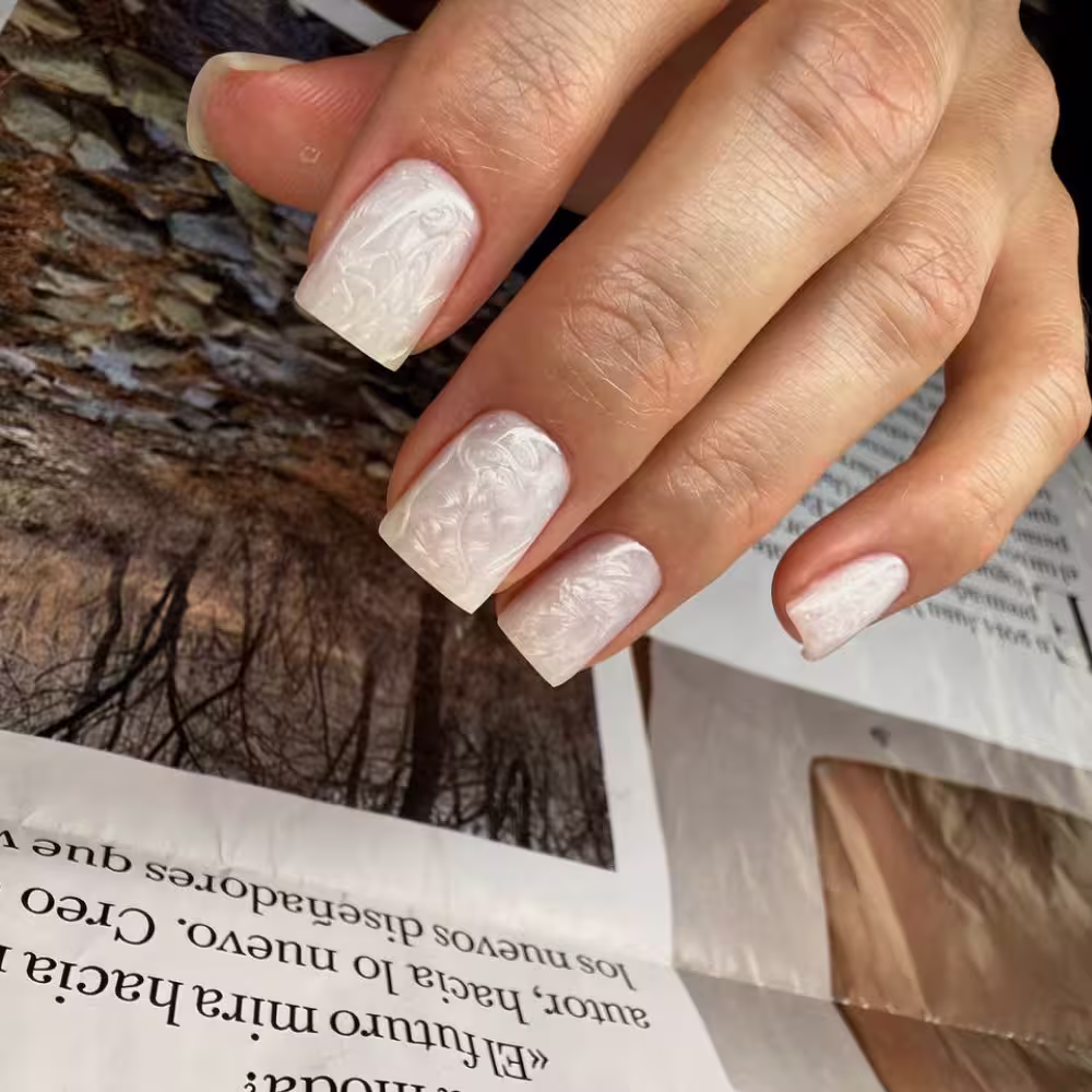 Close-up of a hand with square-shaped nails painted in white with embossed floral designs.