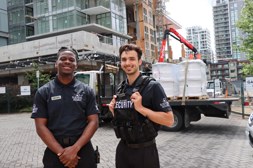 Two security personnel standing in front of a truck with construction equipment in an urban setting.