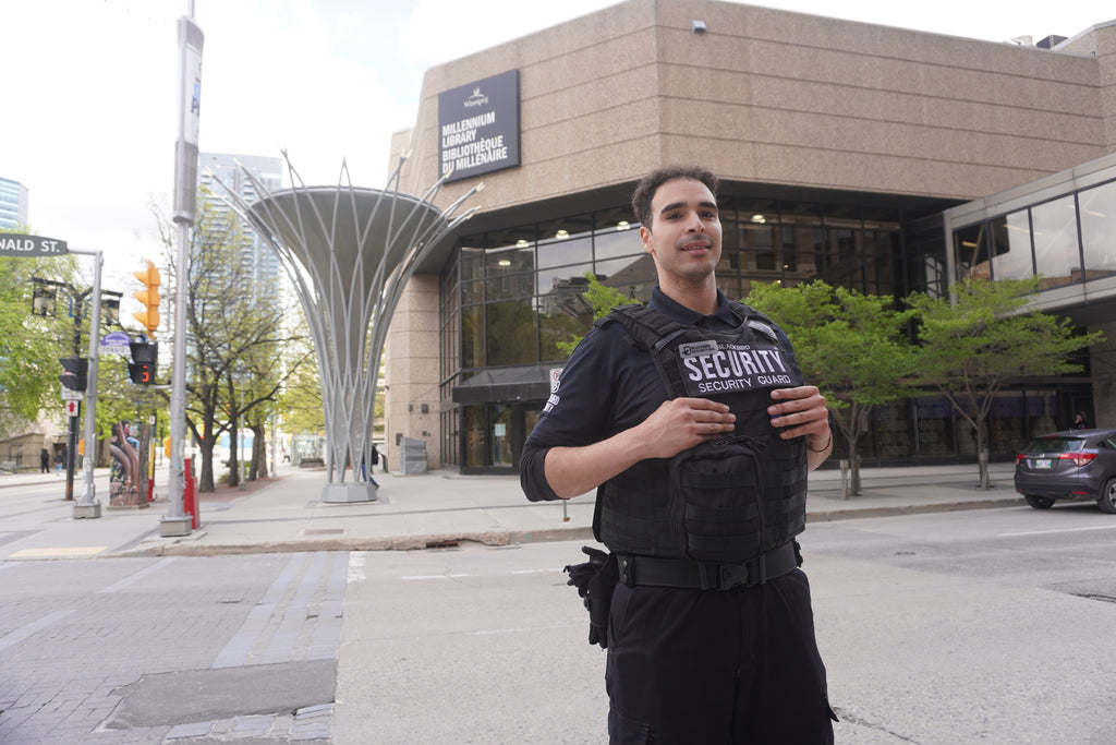A Blackbird Security guard wearing a tactical vest over his uniform stands in front of the Winnipeg Millennium Library.