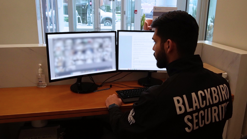 A security guard sits in an office chair at a front desk, monitoring CCTV footage displayed on the screens. The back of his jacket is emblazoned with the Blackbird Security logo.