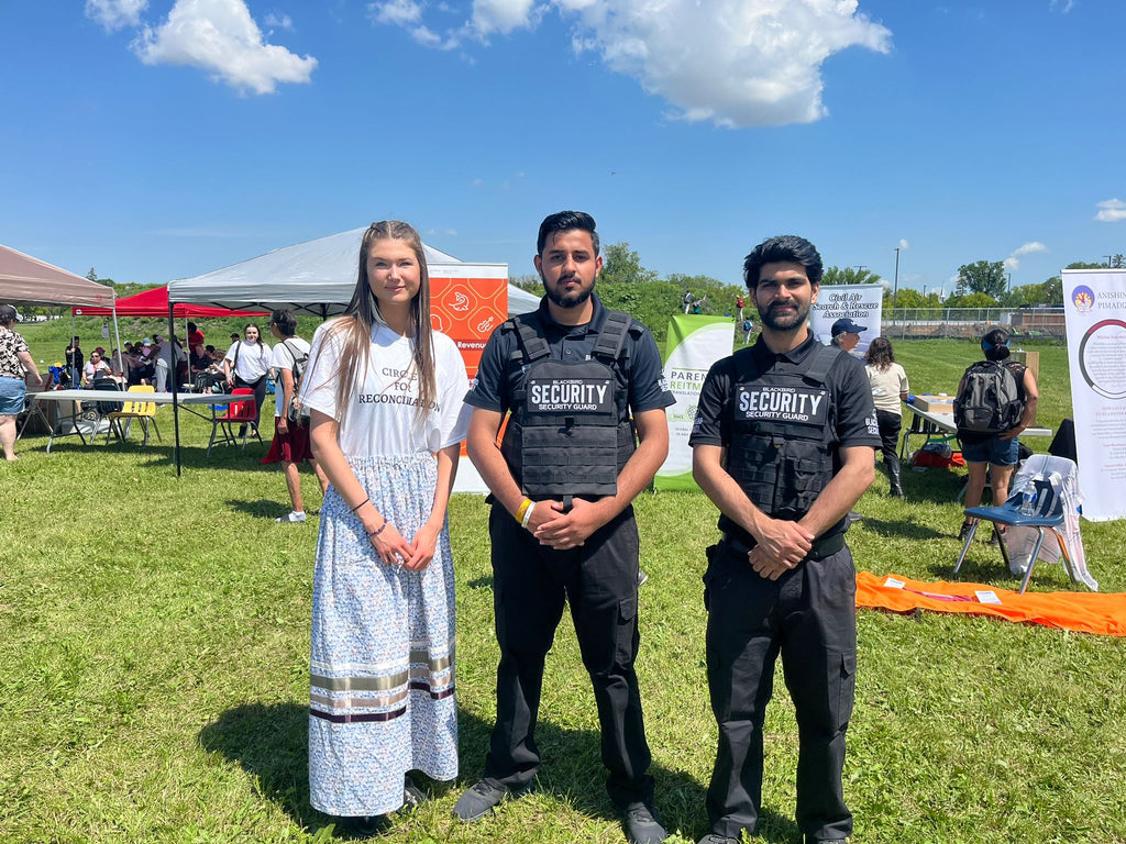 Two Blackbird Security event security guards stand beside a woman wearing a white t-shirt and skirt at the Circles for Reconciliation event in Winnipeg, Canada.