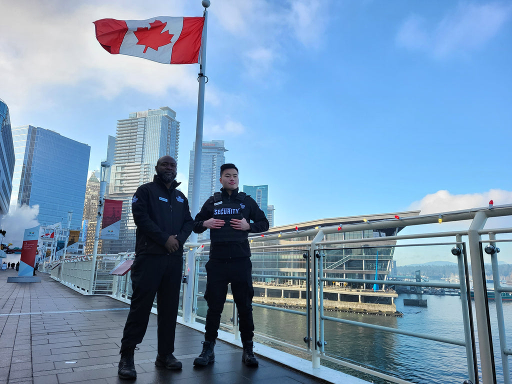 Two Blackbird Security guards stand at Vancouver's Canada Place convention centre, overlooking the water. A Canadian flag flies in the background.