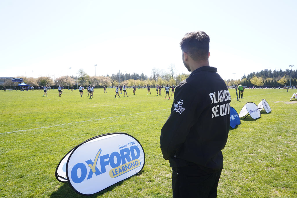 An event security guard stands with his back to the camera, surveying grass sports field at UBC. In the background, a game of rugby is played between teams of women wearing white and black jerseys. The back of the guard's black uniform jacket reads "Blackbird Security".