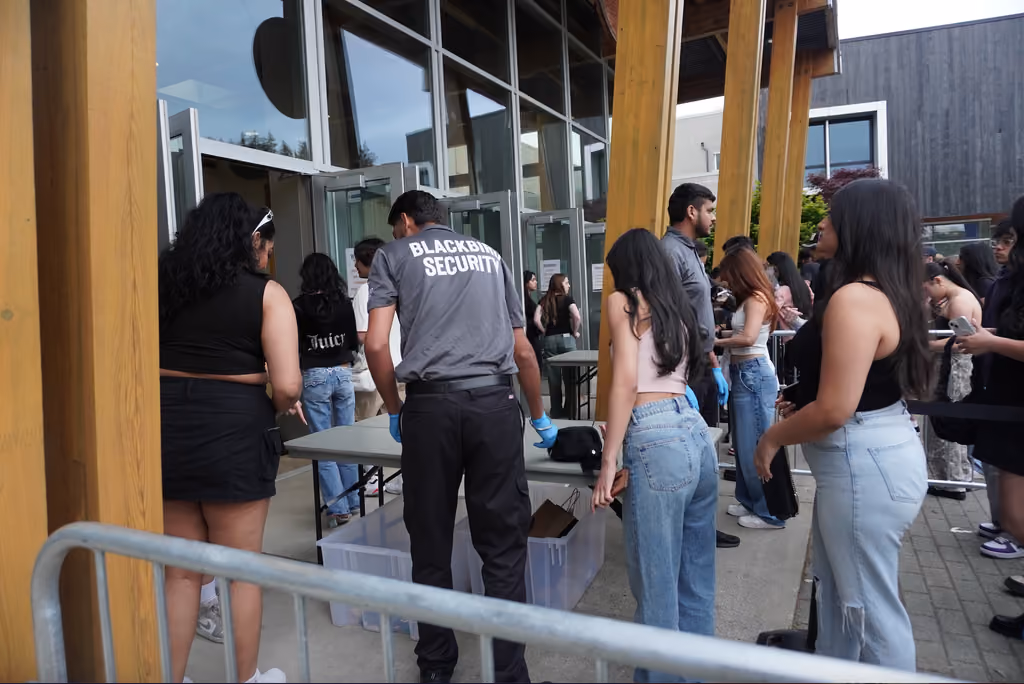 Event security guards from Blackbird Security conduct bag checks at a venue in Vancouver, Canada. The shot contains a security guard with his back to the camera, as well as orderly lines of concert attendees.