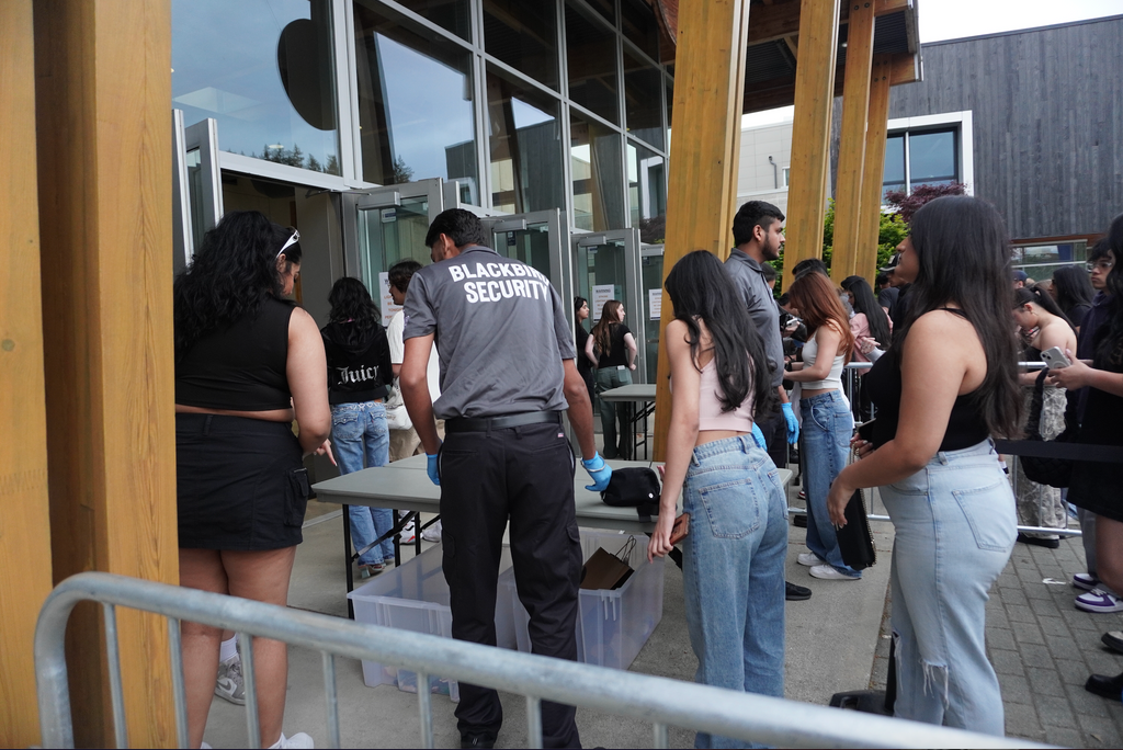 Event security guards from Blackbird Security conduct bag checks at a venue in Vancouver, Canada. The shot contains a security guard with his back to the camera, as well as orderly lines of concert attendees.