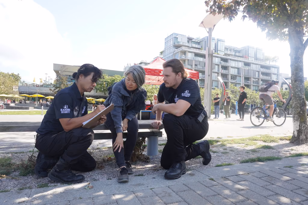 Two Blackbird Security guards kneel on the ground helping a woman sitting on a bench who has apparently injured her leg. The female security guard on the left is making a report on a clipboard.