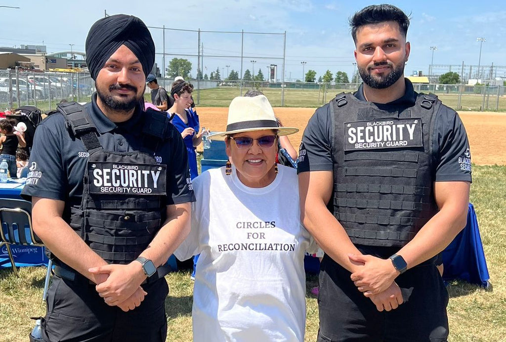 Two Blackbird Security guards in uniform standing on either side of an Indigenous elder wearing a white t-shirt that says "Circles for Reconciliation".