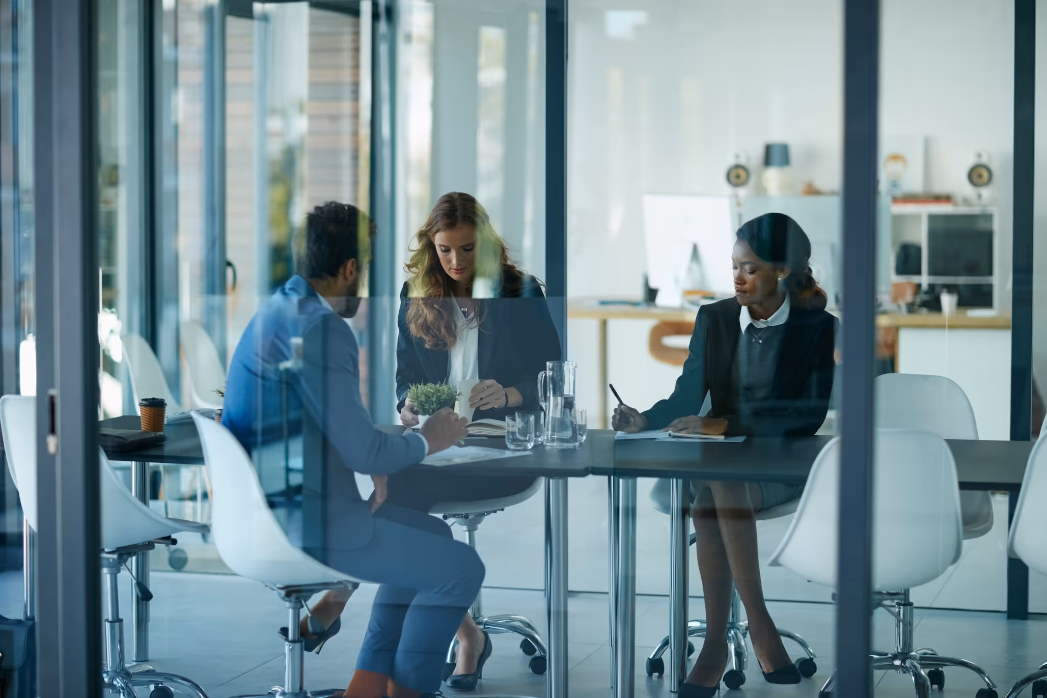 Three business professionals in formal attire having a meeting around a table in a modern glass office room.
