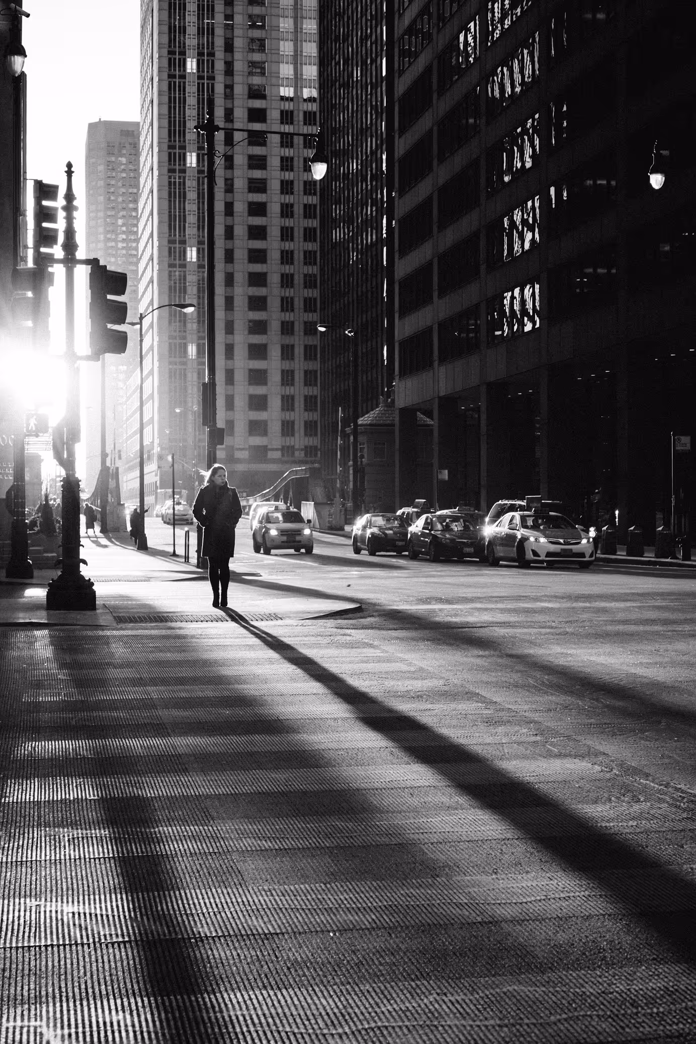 Woman walking alone on a city street at sunset, casting a long shadow with tall buildings and cars in the background.