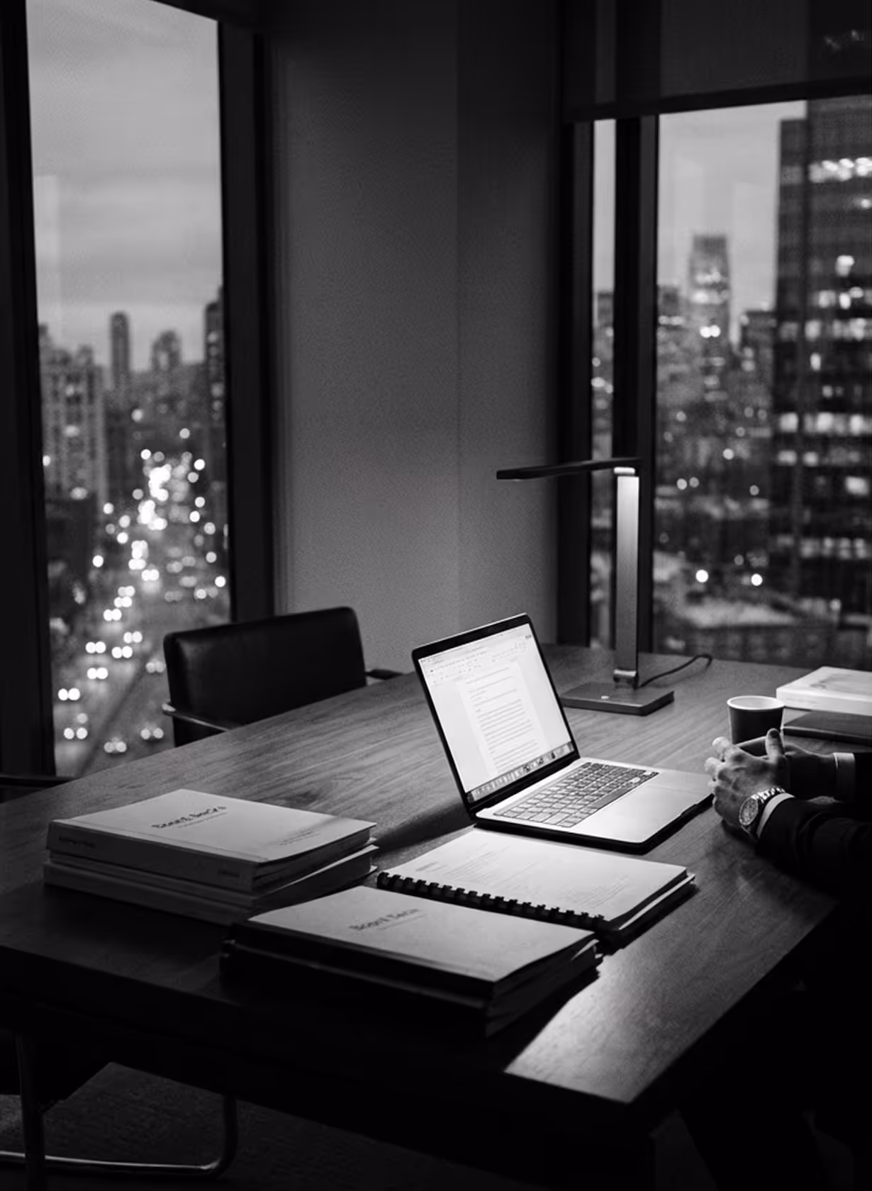 Dimly lit office with a wooden table holding a laptop, stacks of documents, notebook, desk lamp, and a cup, with a cityscape visible through large windows at dusk.