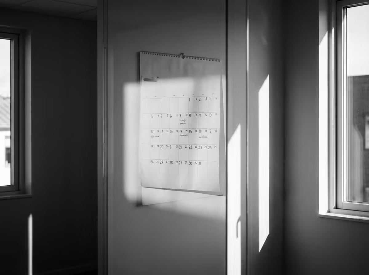 Black and white photo of a monthly calendar hanging on a wall between two windows with sunlight casting shadows.