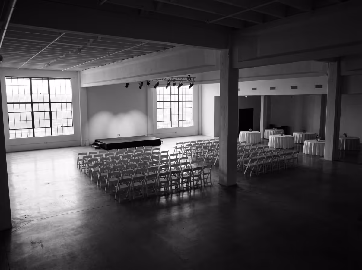 Empty industrial-style event space with rows of folding chairs facing a small stage and round tables with tablecloths in the background.