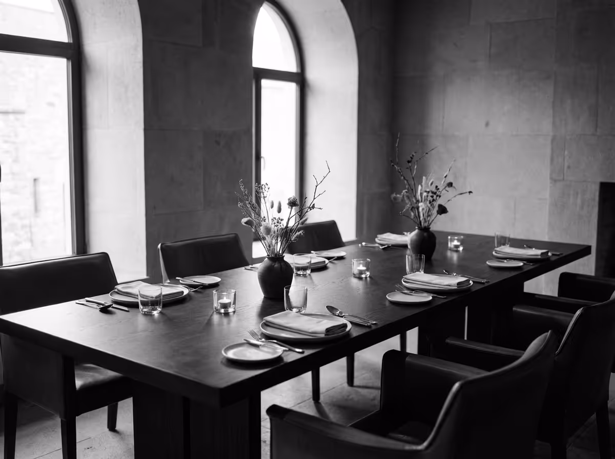 Black and white photo of a dining table set with plates, glasses, cutlery, candles, and two vases with dried flowers in a room with arched windows.