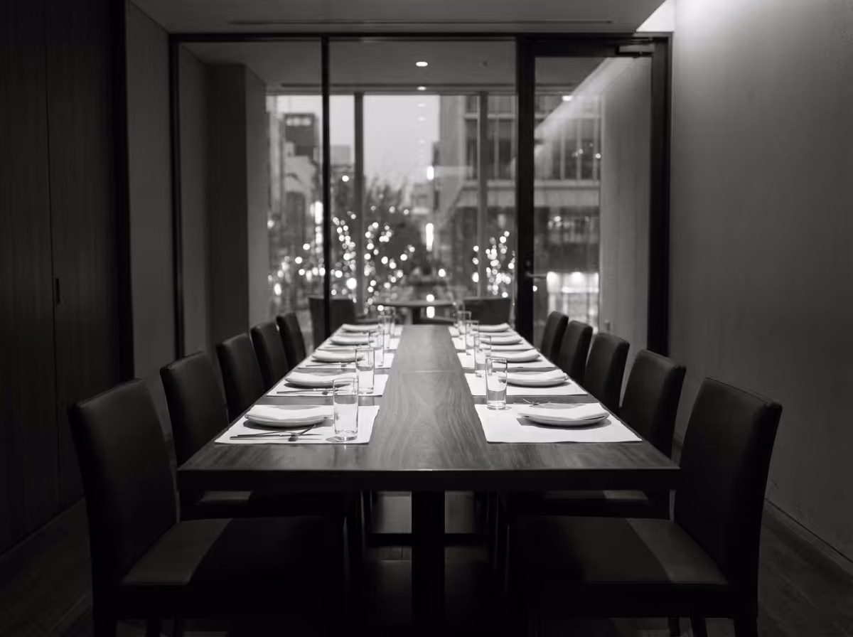 Long dining table set with plates, glasses, and cutlery in a modern room with large windows showing city lights outside.