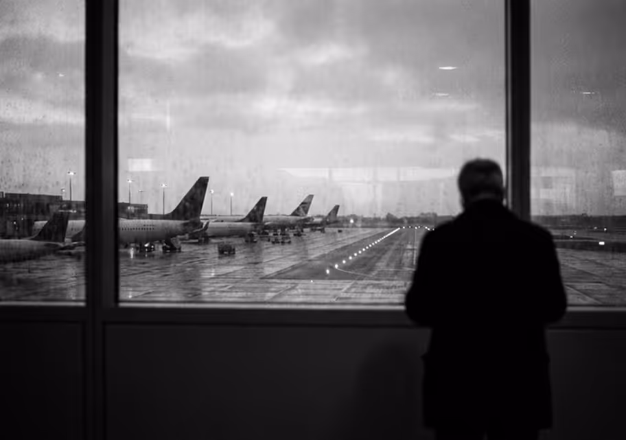 Silhouette of a person looking out a large window at an airport runway with multiple parked airplanes on a rainy day.