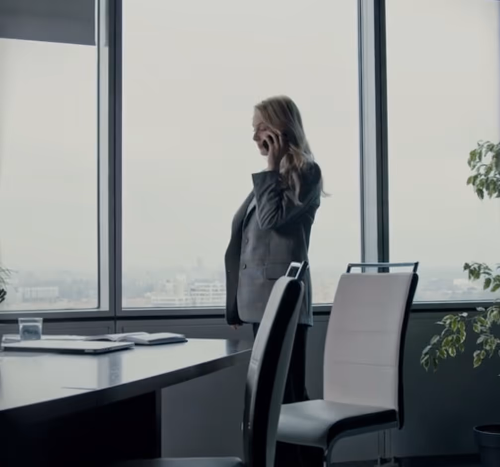 Woman in a gray blazer talking on the phone in a modern office with large windows and a city view.