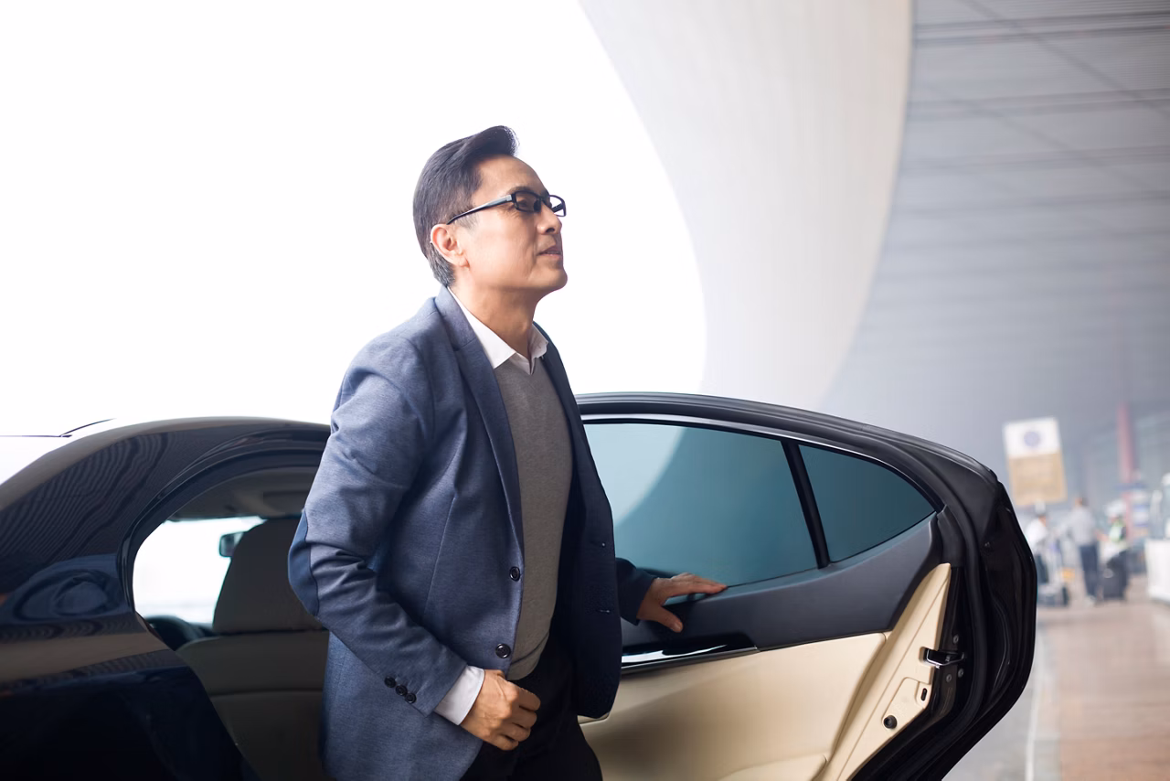 Man in glasses and business attire exiting a black car at an airport or similar transport hub.