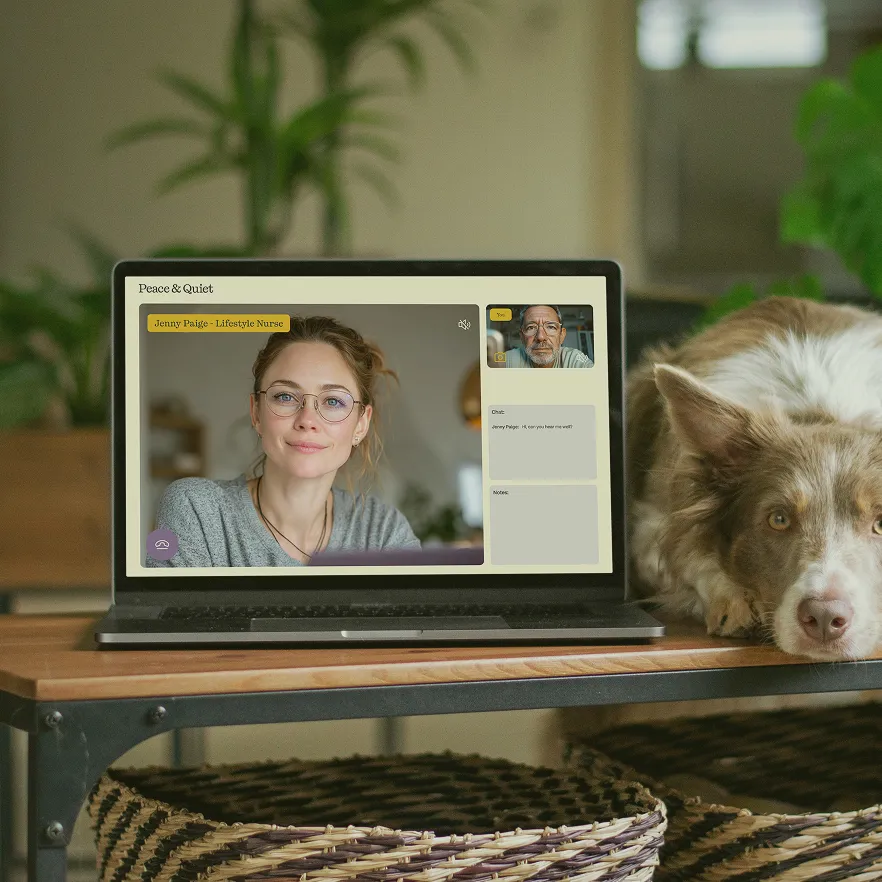 Laptop on wooden table showing a video call between a woman labeled Jenny Paige - Lifestyle Nurse and an older man, with a dog resting its head on the table beside the laptop.