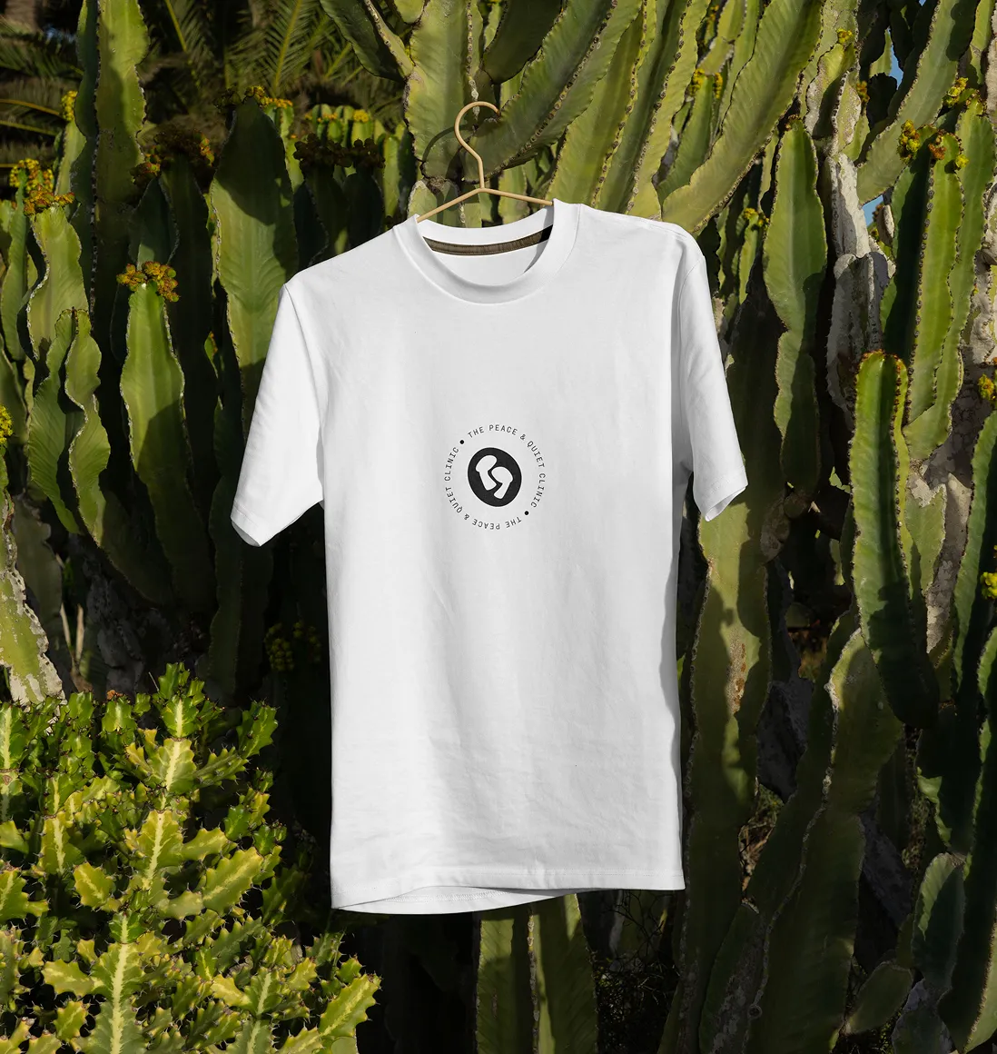 White T-shirt with a circular black logo and text that reads 'The Peace & Quiet Clinic' hanging on a wooden hanger in front of tall green cactus plants.
