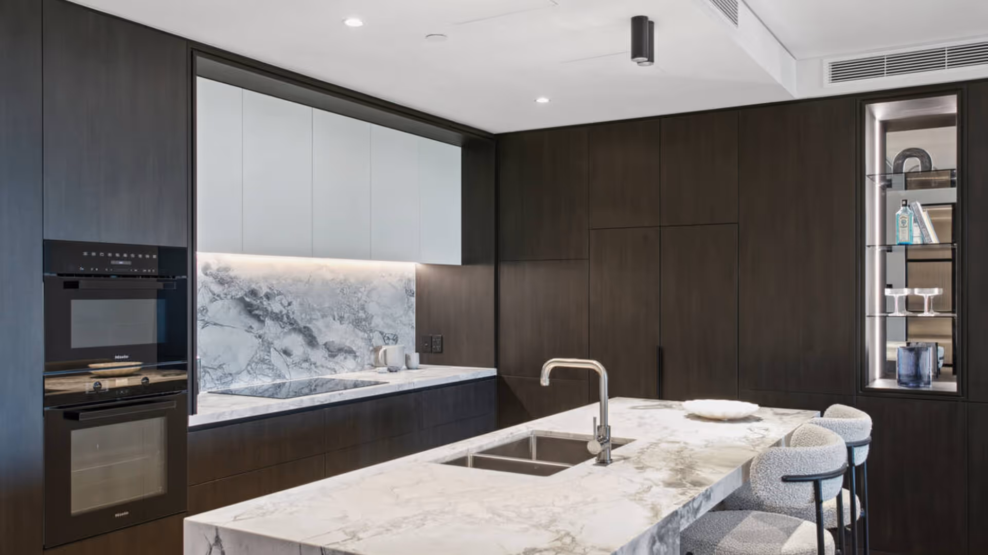 Modern kitchen with dark cabinetry, gray marble island with three black bar stools, and a vase of greenery on the countertop.